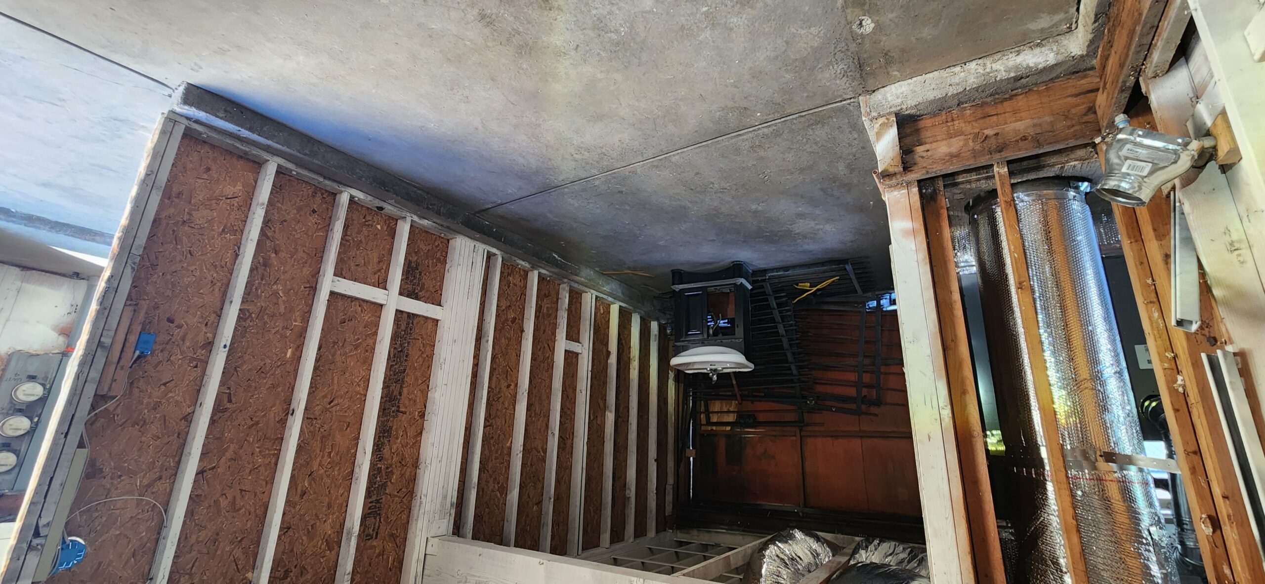 Partially constructed room with exposed wooden studs, visible wiring, ductwork, and a concrete ceiling. A light fixture and other construction materials are present.