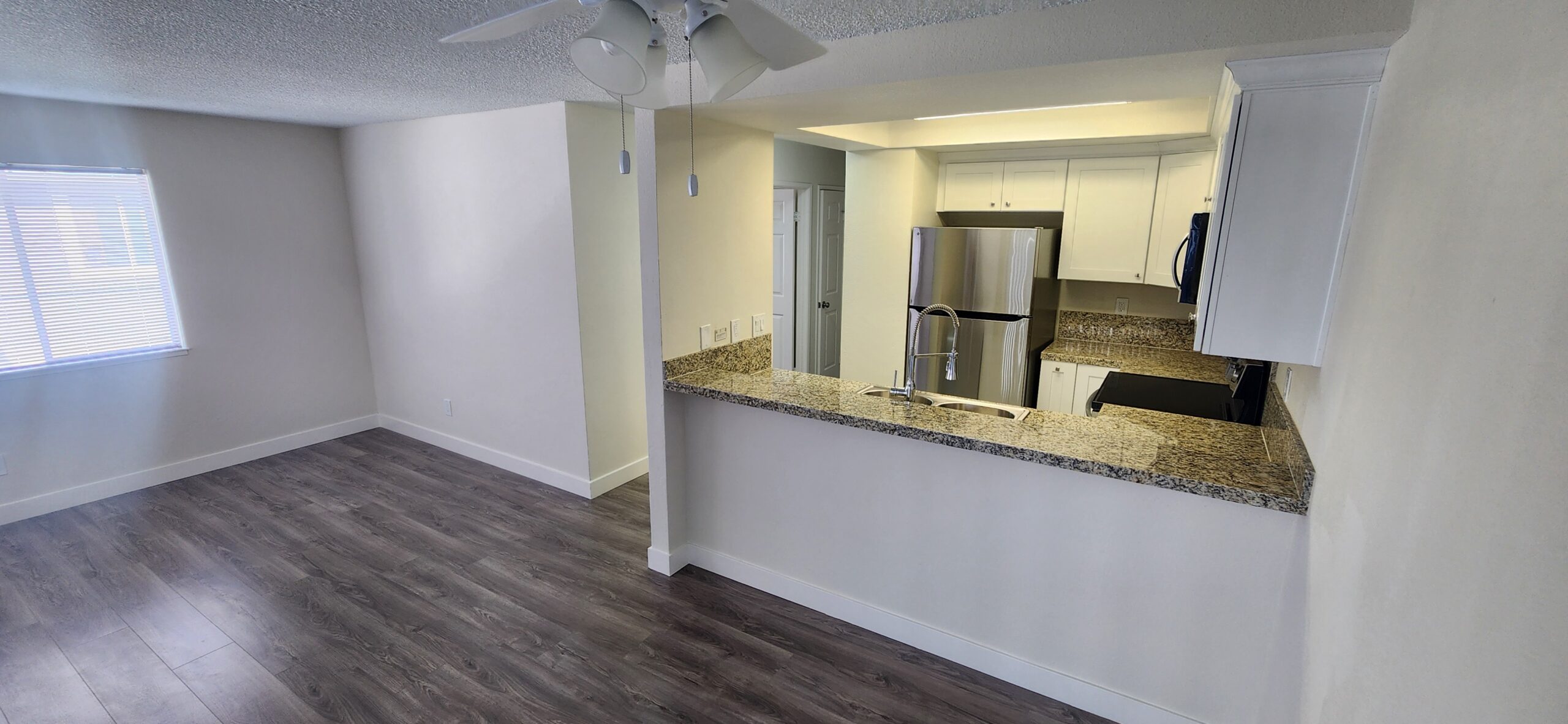 Modern apartment with wood floors, an open living area, ceiling fan, and a kitchen featuring granite countertops, stainless steel appliances, and white cabinets.